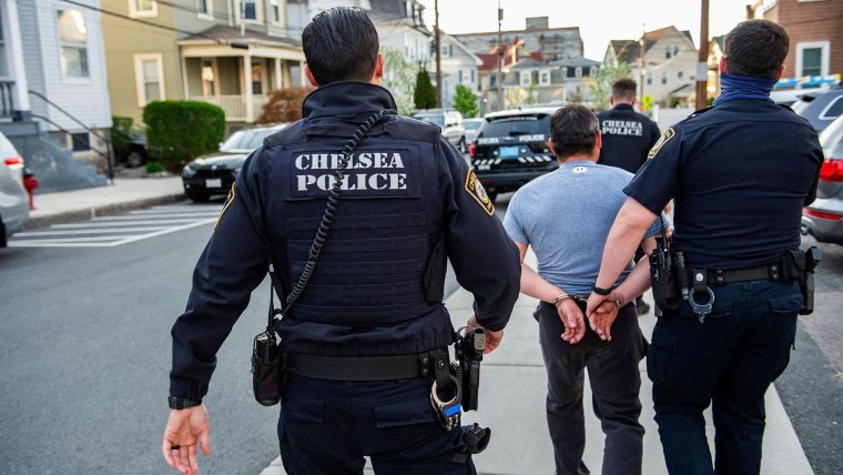 Tres policías armados con chalecos antibalas que dicen "Chelsea Police" escoltan a un hombre latino que está esposado y dando la espalda a la cámara por una calla de Chelsea, Massachusetts, en mayo de 2021