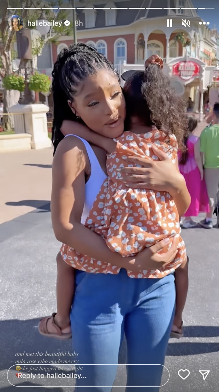 halle bailey a 22 year old black woman with braids in a ponytail in a white tank top and jeans holds a young girl of color named mila rose, wearing an orange and white floral dress, tan sandals, and Minnie ears at disney world