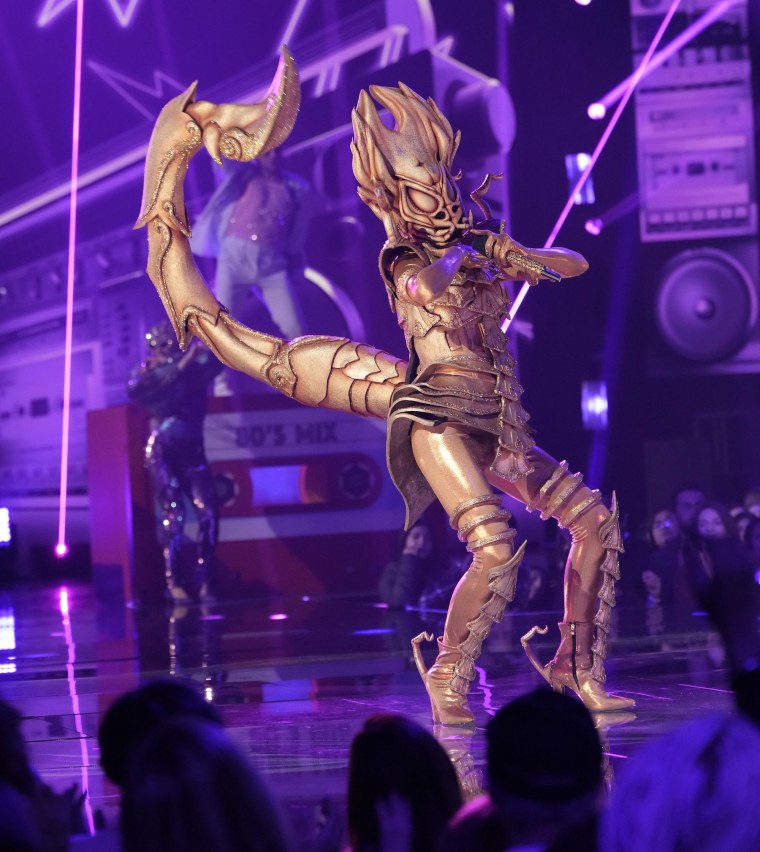 A person in a gold sparkly scorpion costume stands on stage holding a mic against a purple backdrop.
