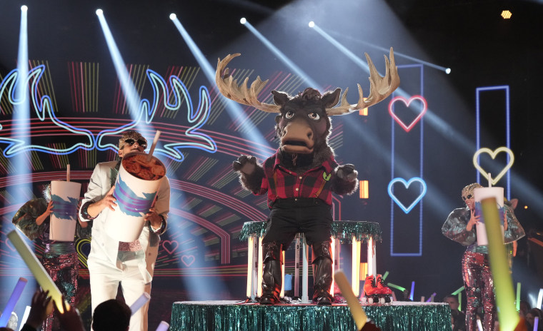 A person in a moose costume stands on stage as dancers run around him holding large sodas