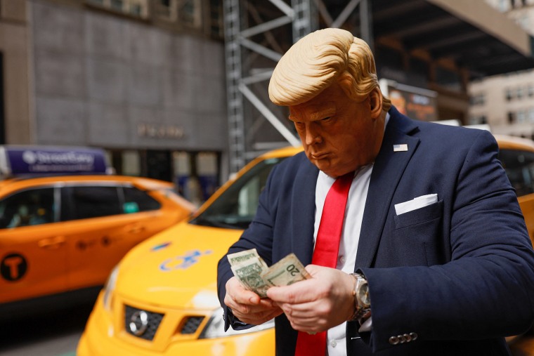 Una persona con una máscara que representa al expresidente Donald Trump, cuenta dinero afuera de la Torre Trump.