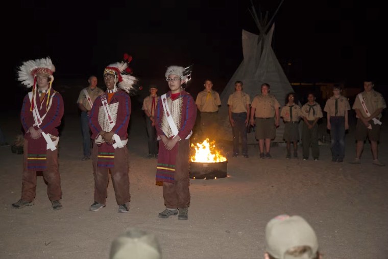 Order of the Arrow nominees stand in line, background, as three members of the order, dressed in Native American regalia, explain what the nomination of the scouts behind them signifies during their Call Out Ceremony at Camp Wilson during the Boy Scout Camp Out for local Boy Scouts of America troops in 2016.