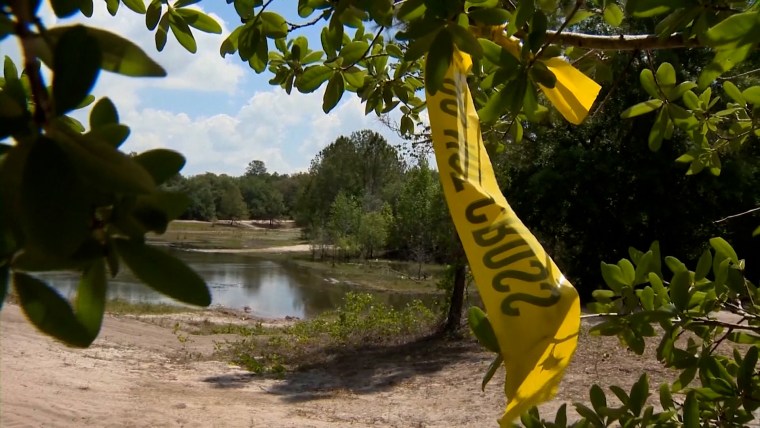 Yellow police tape blows in the wind near the camera in front of a small pond.