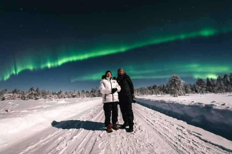 Two women in winter clothes on a snowy background stand in front of green glowing Northern Lights