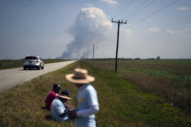 Smoke, reportedly from a burning sugar cane field, rises in the background as farm workers and their allies take a break during a five-day march to push for better wages and working conditions on March 14, 2023, in Pahokee, Fla. 