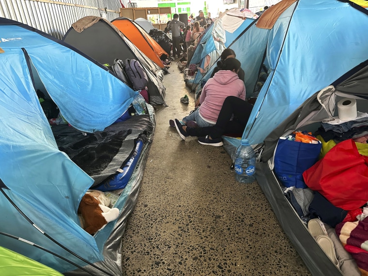 Teresa Muñoz, seated at center wearing a pink hoodie, rests at a tent at a migrant shelter where she has been trying unsuccessfully for about a month to get an appointment to enter the United States through a new U.S. government mobile phone app in Tijuana, Mexico, Thursday, May 11, 2023. She fled her home in the Mexican state of Michoacan after her husband was killed and she was badly beaten. (AP Photo/Elliot Spagat)