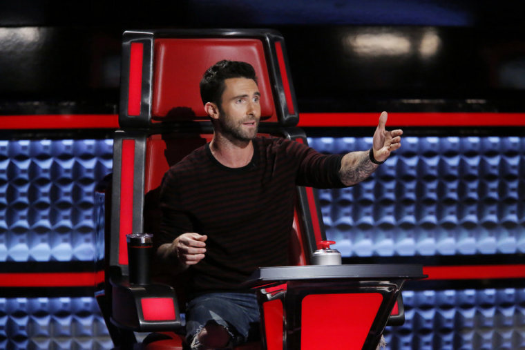 Adam Levine in a red chair on the set of "The Voice" gestures to the right (his left).