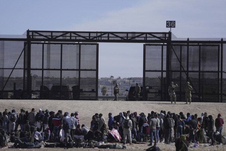 Migrantes que cruzaron la frontera de México a EE.UU. esperan junto al muro fronterizo estadounidense, donde agentes de la Patrulla Fronteriza montan guardia.