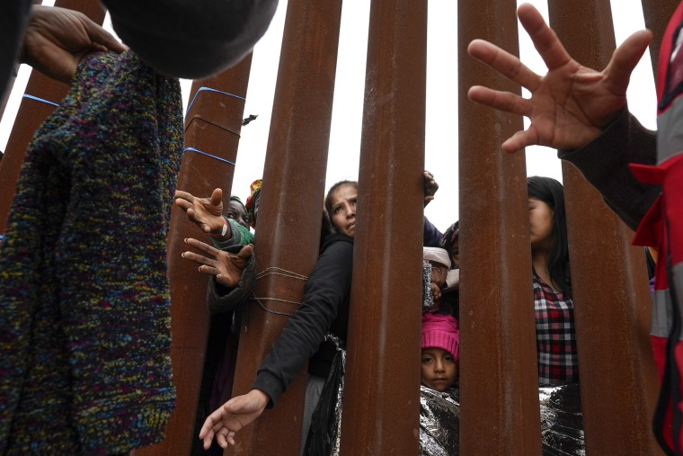 Varias personas extienden sus brazos a través del muro fronterizo para recibir ropa que entregan voluntarios, mientras esperan para aplicar por asilo en San Diego, California, este 12 de mayo de 2023. 