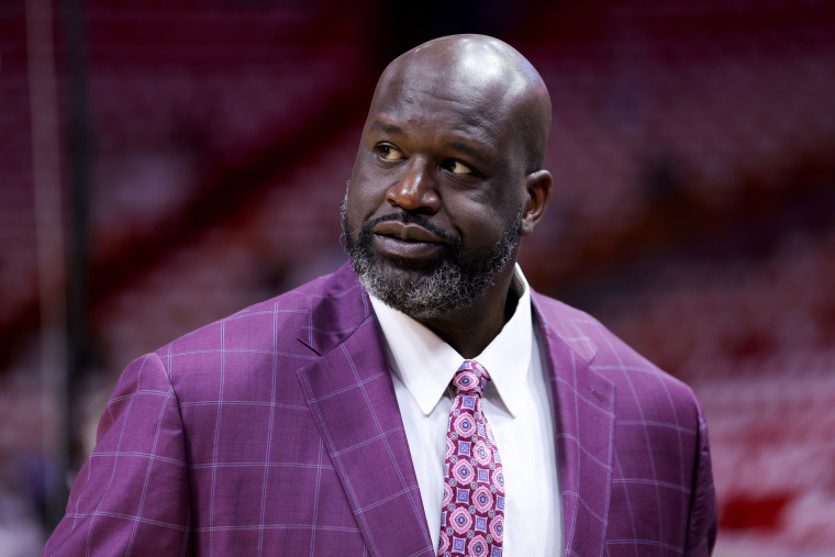 Shaquille O'Neal looks to his right over his shoulder in a basketball arena. He's wearing a purple plaid suit and coordinating tie.