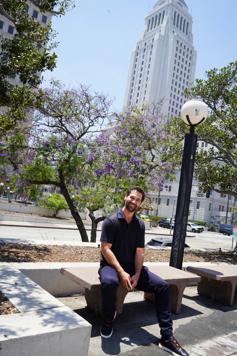 Manuel Garcia-Rulfo sits on a bench in Downtown Los Angeles in front of a flowering purple tree and white art deco building