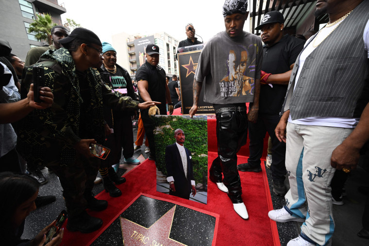 Un groupe d'hommes se rassemblent autour d'une star du Hollywood Walk of Fame installée sur un tapis rouge.  Ils tiennent un portrait du regretté rappeur américain Tupac Shakur.