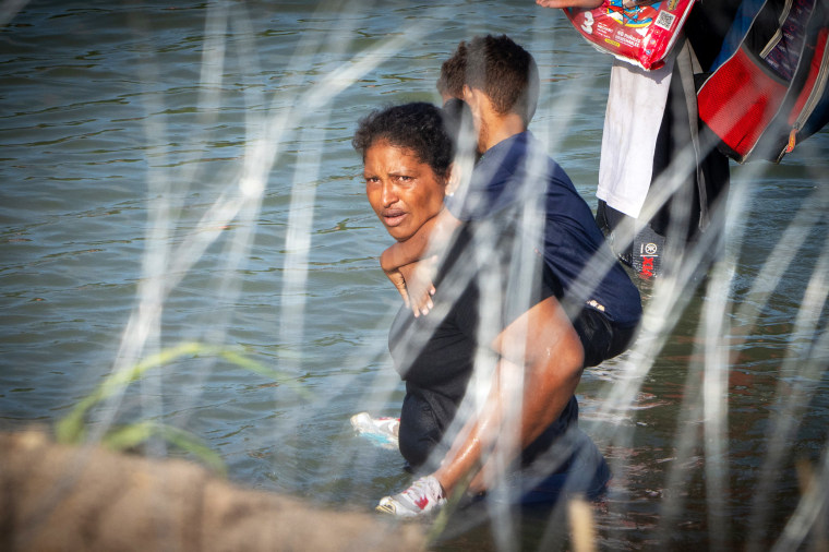 TOPSHOT - A woman holds a child on her back as migrants walk near concertina wire in the water along the Rio Grande border with Mexico in Eagle Pass, Texas, on July 16, 2023. A nearby buoy installation is part of an operation Texas is pursuing to secure its borders, but activists and some legislators say Governor Greg Abbott is exceeding his authority.