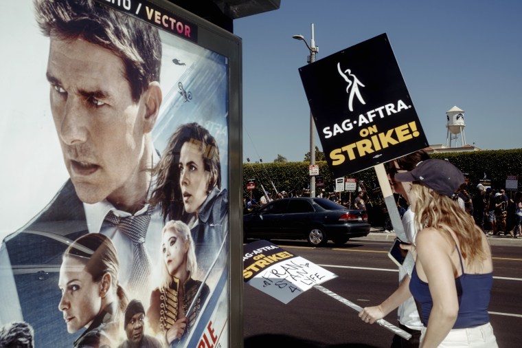 Image: Screen Actors Guild and Writers Guild of America members walk the picket line next to a poster for the movie "Mission Impossible" outside of Paramount Pictures in Los Angeles on July 14. 