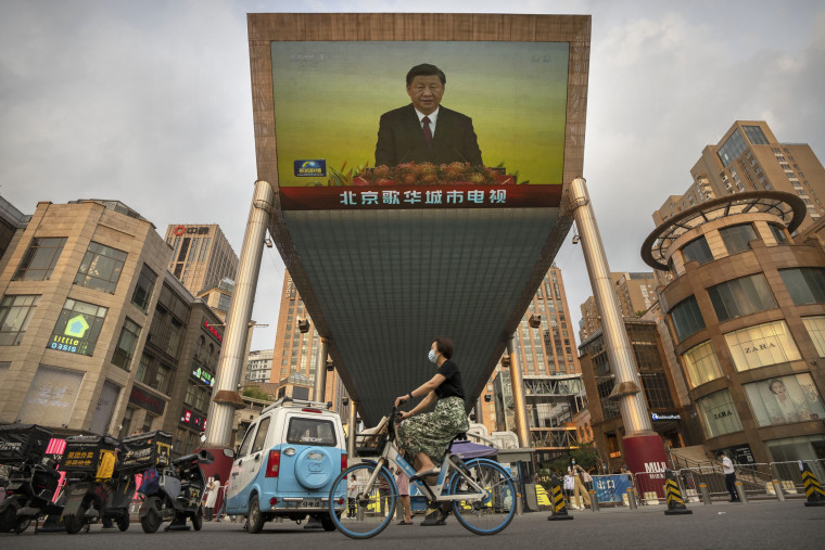 A woman wearing a face mask rides a bicycle past a large television screen at a shopping center displaying Chinese state television news coverage of 