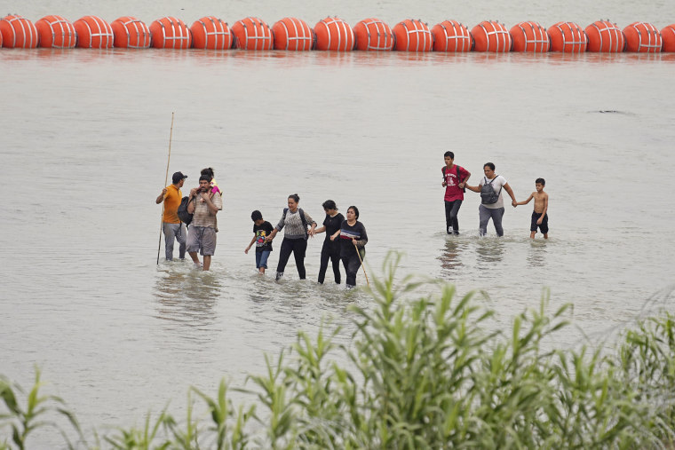 Migrants who crossed the Rio Grande from Mexico walk past large buoys being deployed as a border barrier in Eagle Pass, Texas, on July 12, 2023.