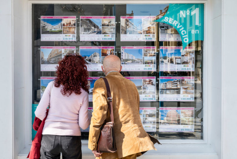 Una pareja observa propiedades a la venta y para alquiler en la vidriera de una oficina de bienes raíces en España.
