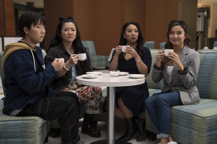 Four people holding white coffee cups look at something offscreen. They are sitting at a table on green couches.
