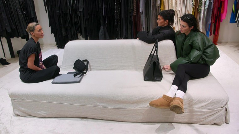 Kim Kardashian in a t-shirt and sweats sits on the left side of a couch looking at Kendall and Kylie Jenner, both in outerwear, on the other side.