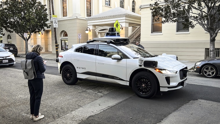 Waymo communications manager Julia Ilina stands next to a Waymo driverless taxi that stopped in the street because the back door was not completely shut, while traffic backed up behind it, in San Francisco, on Feb. 15, 2023. 