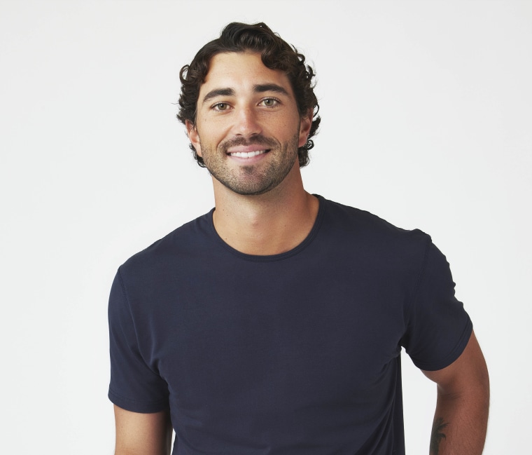 Joey Graziadei smiles in a blue crew neck tshirt against a white background.