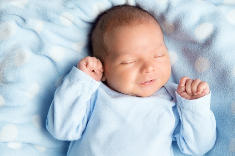 Newborn Baby Sleeping Smiling. Cute Infant Child in Wrap Bodysuit. New Born Little Boy smile in Blue Clothes lying on Blanket. Small Kid Face Close up Portrait holding Hands First