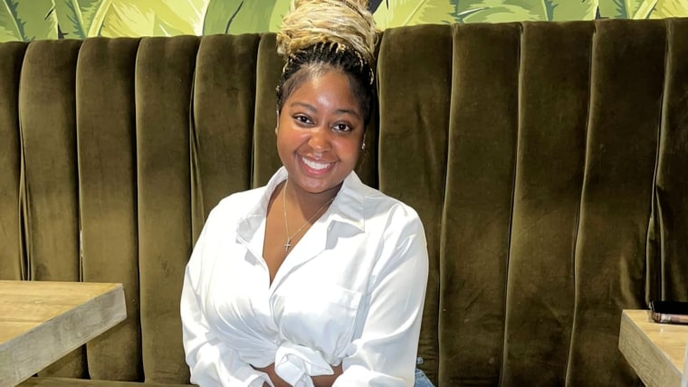 A young Black woman with her hair pulled into a bun (blond braids with dark roots) smiles on a green velvet couch.sShe's wearing a cross necklace and a white button down shirt.