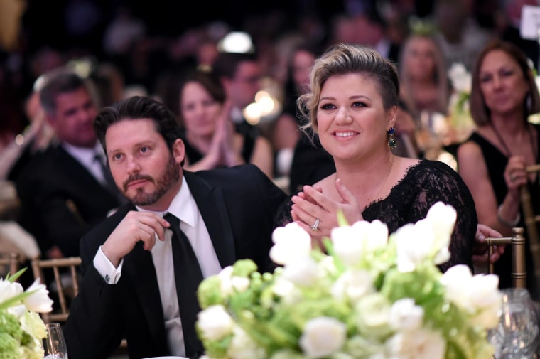 Kelly Clarkson and Brandon Blackstock in formal attire sit at a table. White flowers can be seen in the foreground.