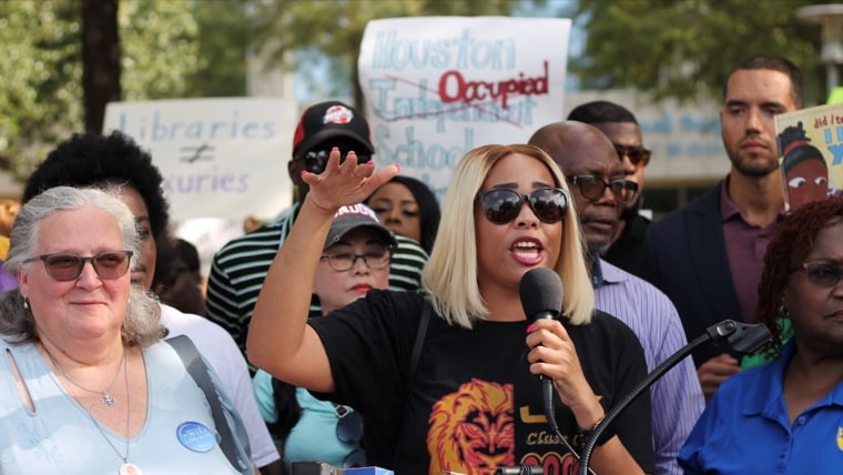 Una protesta reciente de padres de familia que se oponen a los cambios en el distrito escolar de Houston. Hay personas con carteles que dicen "No a la ocupación del distrito escolar" y está hablando una madre de familia frente a un micrófono.