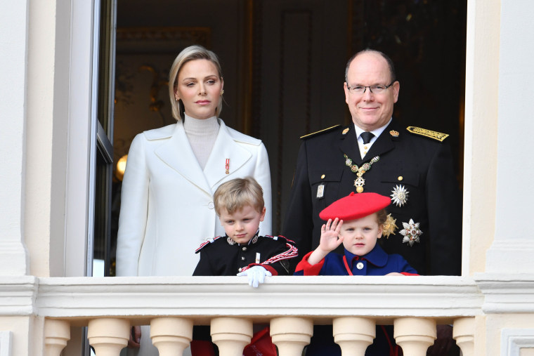 Charlene in a white coat, Albert in a navy regal outfit and their two young children wave from a balcony.