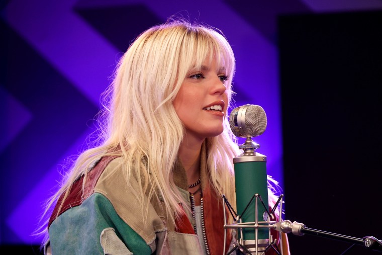 Reneé Rapp in a zip up jacket with bleach blond hair and banks talks into a blue and silver microphone.