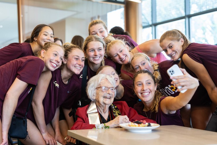 Sister Jean sits at a table surrounded by young women in maroon loyola shirts as they take a selfie.