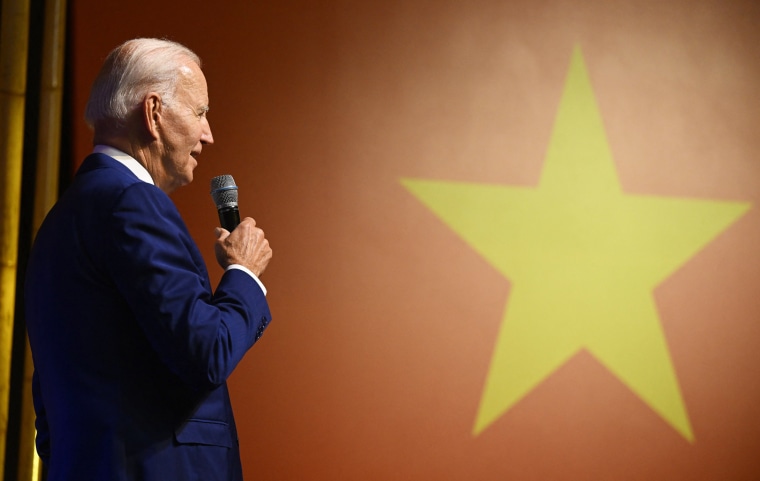 Joe Biden holding microphone in front of Vietnam's flag. 