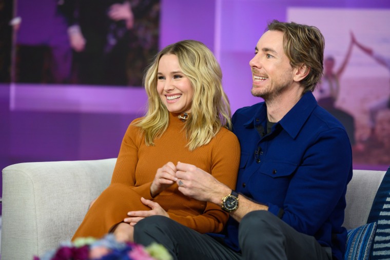 Bell and Shepard sit smiling on a couch on a TV studio set. Bell is in a rust orange turtleneck dress and shepard is in a royal blue shacket.