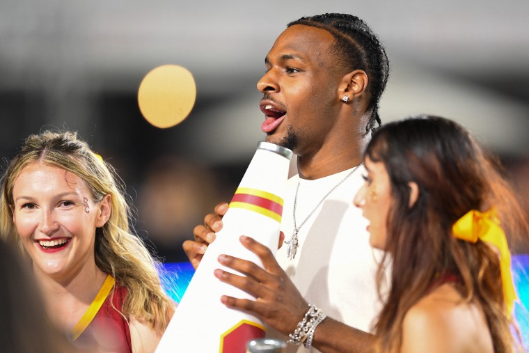 Bronny James holds a white cone with USC logo and shouts at something off-camera. A young blond woman smiles to his right and a brunette on his left looks concerned at something off-camera.