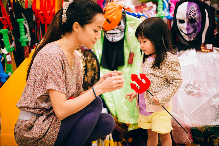 Pretty young mom and lovely toddler girl shopping for Halloween costume accessories in the department store.