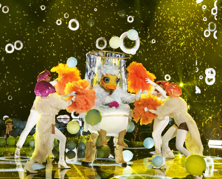 A person in a rubber duck costume in a bathtub dances onstage with four dancers in shower caps