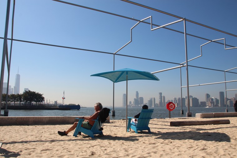 Two people sit in blue beach chairs on the sand of Gansevoort Peninsula beach, overlooking the Hudson River in Manhattan