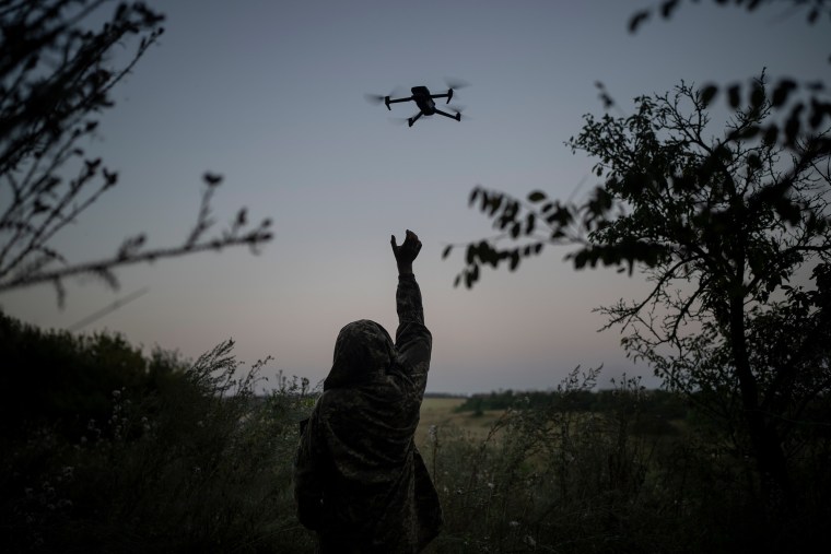 A drone pilot, seen from the back, reaches for a drone in a field.