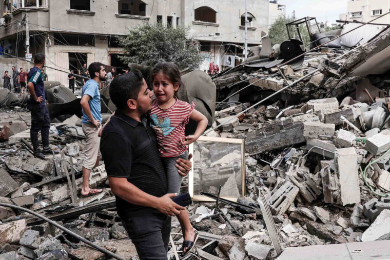 Image: A man carries a crying child past a destroyed building in Gaza City.