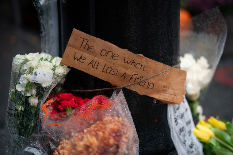 Image: A condolence sign surrounded by flowers reads "The one where we all lost a friend."
