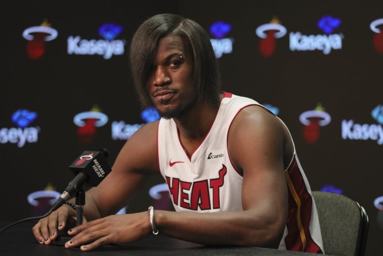 Butler in a Miami Heat jersey speaks into a microphone. His hair has been straightened and his bangs cover his right eye. He also has several facial piercings.