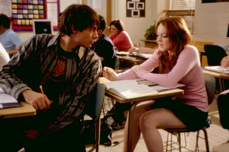 Jonathan Bennett in a striped button down and graphic tee looks over his left shoulder while sitting at a desk. Lindsay Lohan responds behind him, also seated at a desk wearing a pink sweater.