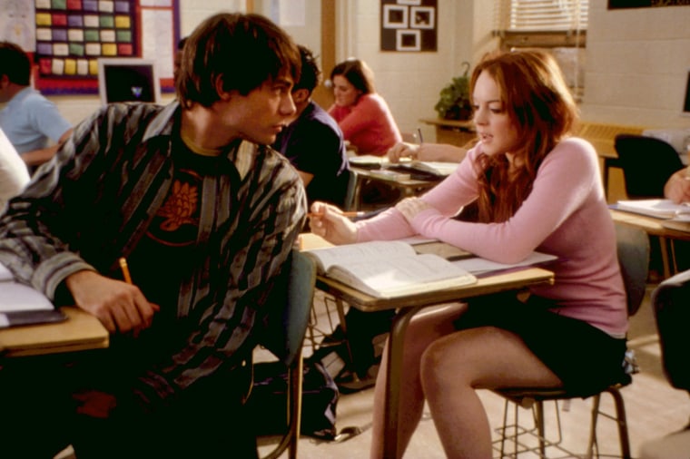 Jonathan Bennett in a striped button-down and graphic T-shirt looks over his left shoulder as he sits at a desk. Lindsay Lohan responds behind him, also sitting at a desk in a pink sweater.