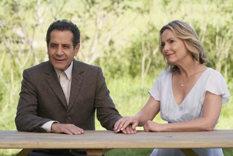 Shalhoub as Adrian Monk in a brown suit sits outside and holds hands with Melora Hardin as Trudy, who is wearing a white dress with a v-neck and flutter sleeves.