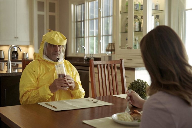 Shalhoub as Monk sits at a kitchen table in a yellow hazmat suit, sipping an iced coffee from a tumbler with a straw in a hole through his suit mask.