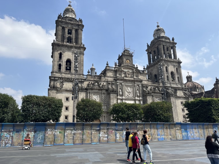 People walk past a metal wall covered in graffiti, blocking the Metropolitan Cathedral