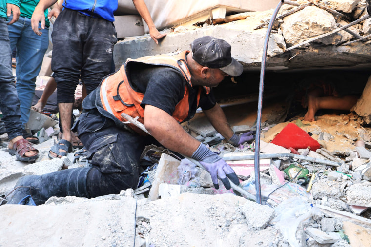 A man in an orange emergency vest kneels and speaks to a person underneath building rubble