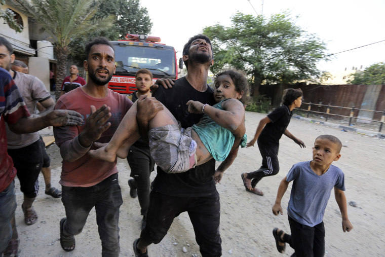 A man, surrounded by other people, carries a girl who is covered in dirt from building rubble along a road