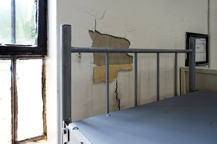 Missing plaster from the wall behind a bunk bed in a single-level housing unit.